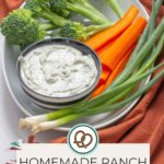 A bowl of creamy homemade ranch dip is nestled on a plate surrounded by vibrant broccoli, crisp carrot sticks, and fresh green onions. The text reads: "Homemade Ranch Dip by happysnackcidents.com.