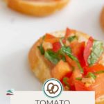 A detailed close-up of a savory tomato bruschetta on a slice of baguette, garnished with fresh herbs. A label elegantly states "Tomato Bruschetta" and features a website link below for more delicious details.