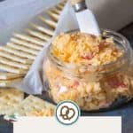 A glass jar brimming with creamy pimento cheese sits beside a spreader, encircled by crispy crackers. The label proudly displays "Pimento Cheese" and happysnackcidents.com.