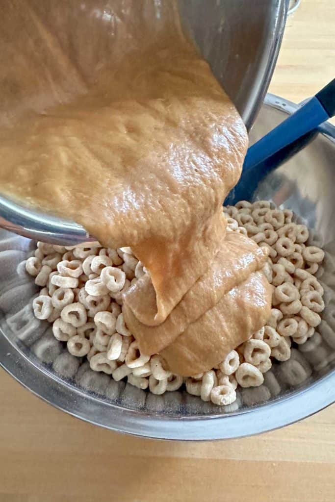 Pouring warmed honey and peanut butter mixture over Cheerios in a bowl.