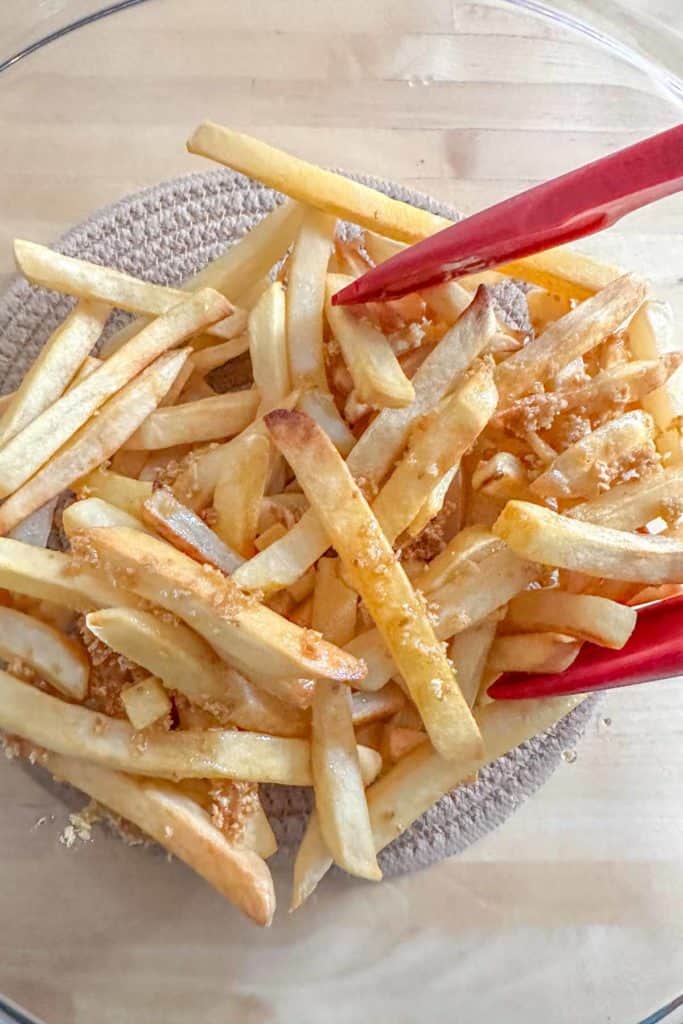 Mixing fries with chopped garlic in a glass mixing bowl using tongs.