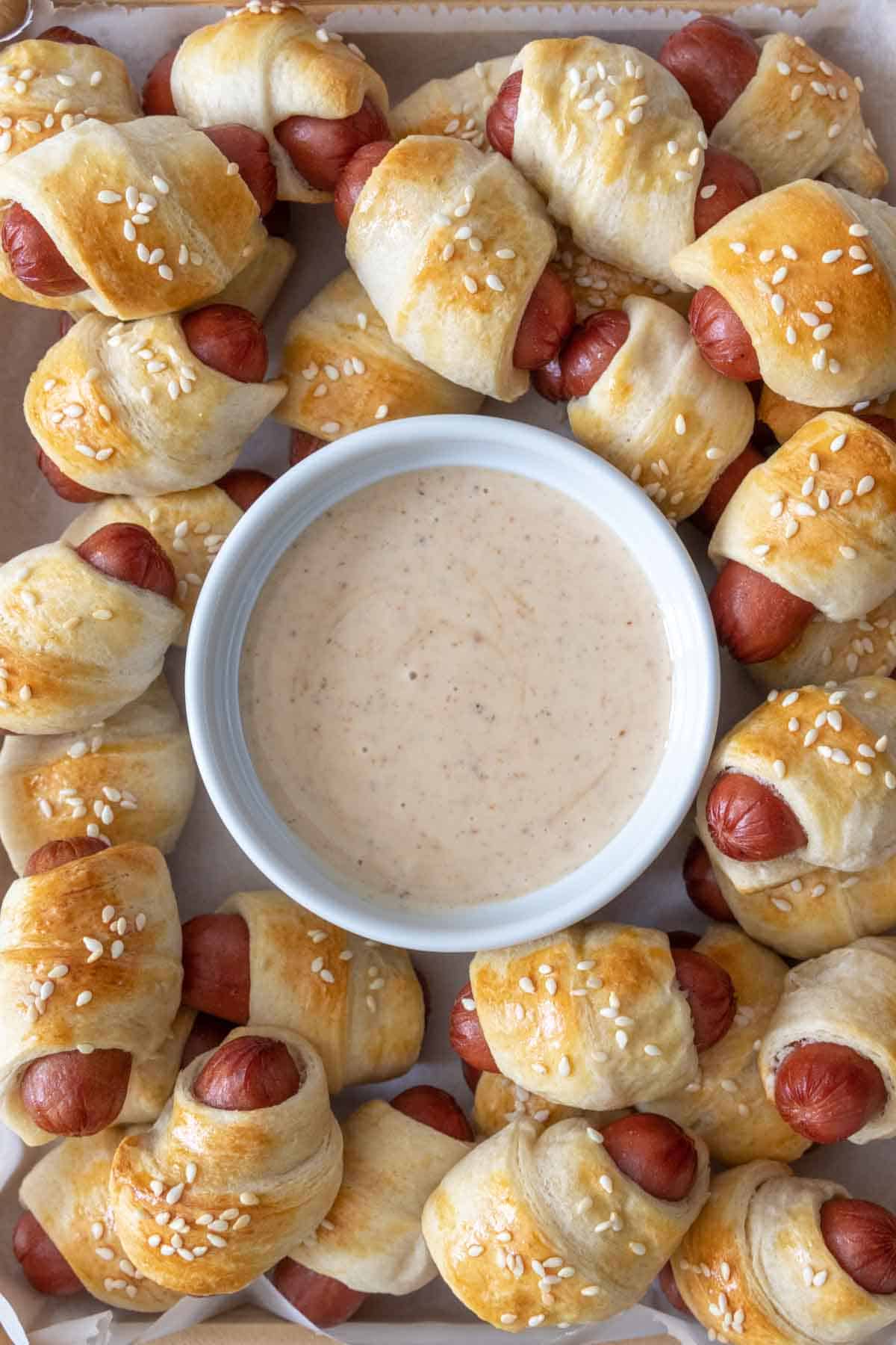 Bowl of sauce surrounded by mini pigs in a blanket appetizers.