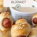 A close-up of pigs in a blanket—classic mini sausages wrapped in golden pastry with sesame seeds—served alongside a small bowl of dipping sauce.