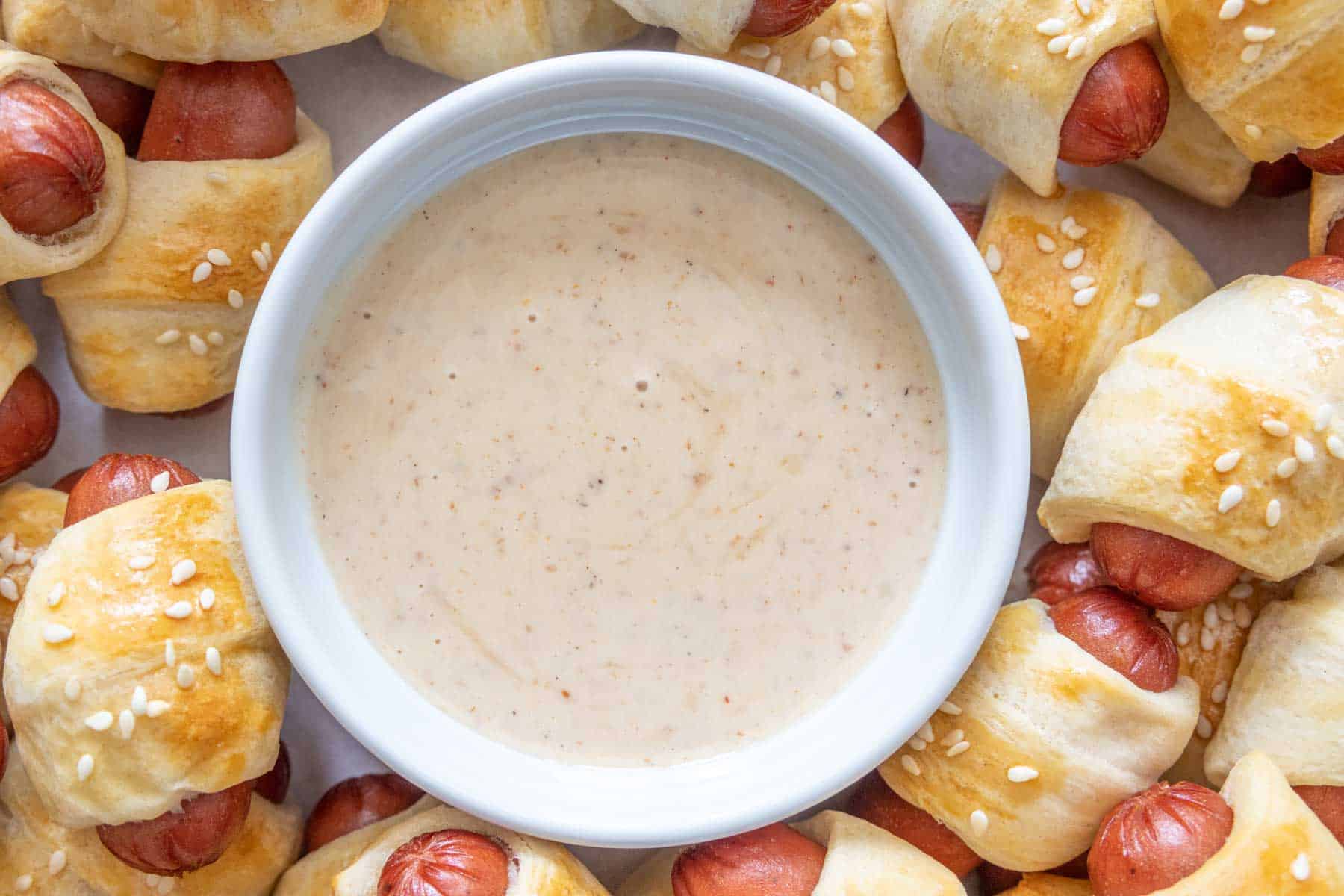White bowl of dipping sauce served alongside pigs in a blanket.
