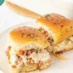 Two chicken bacon ranch sliders sit on a white plate, stuffed with creamy chicken, crispy bacon bits, and herbs, all tucked between golden toasted buns.