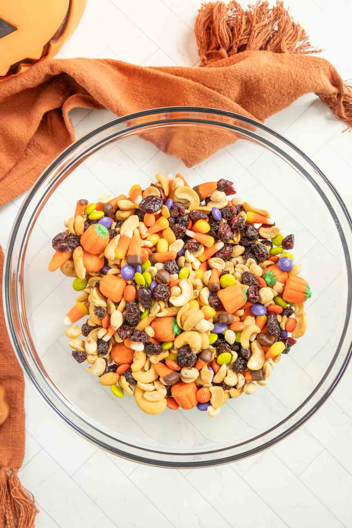 A clear glass bowl filled with a variety of trail mix ingredients, including nuts, raisins, and colorful candy pieces. An orange fabric is placed nearby on a light-colored surface.