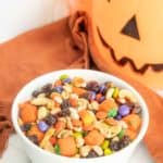 A white bowl filled with a variety of Halloween-themed candies, nuts, and raisins, placed in front of a Jack-o'-lantern decoration.