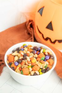 A white bowl filled with a variety of Halloween-themed candies, nuts, and raisins, placed in front of a Jack-o'-lantern decoration.