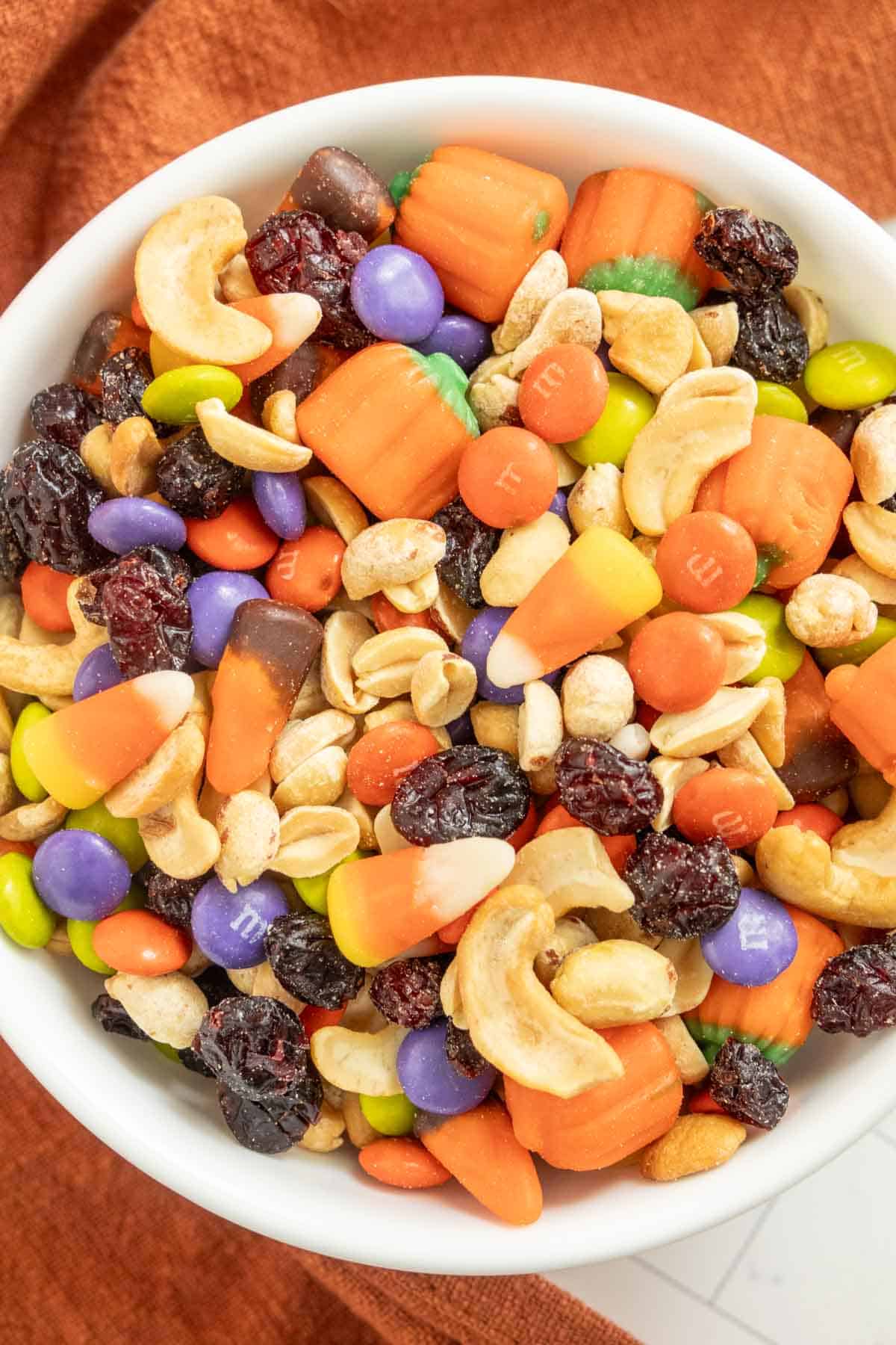 A bowl filled with a colorful assortment of Halloween-themed trail mix, including candy pumpkins, candy corn, chocolate candies, nuts, and dried fruits.