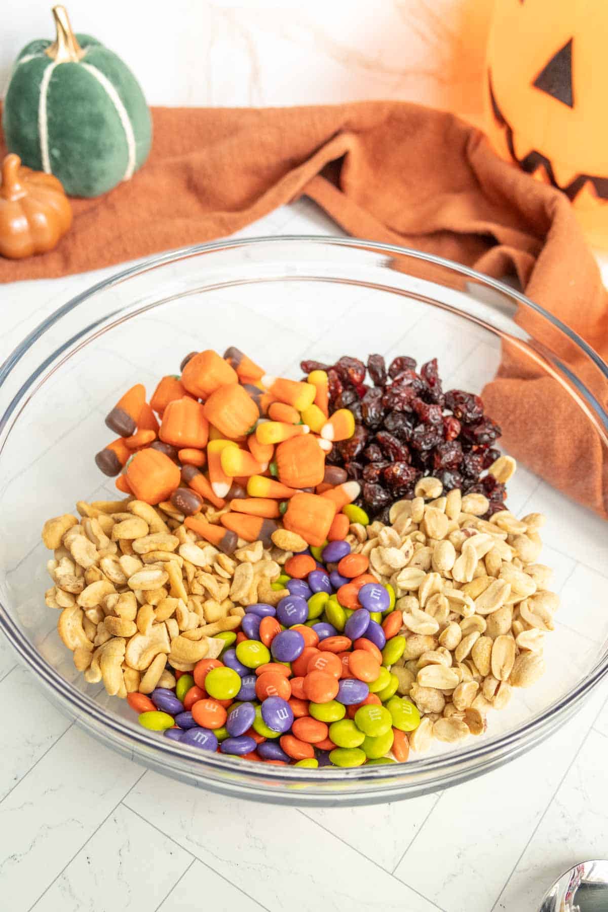 A clear bowl containing peanuts, candy corn, chocolate candies, cashews, and dried cranberries. A pumpkin decoration and an orange cloth are in the background.