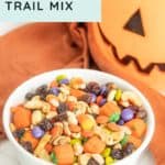 A white bowl of halloween trail mix, filled with candy corn, raisins, peanuts, and candy pieces, sits next to a plastic jack-o'-lantern and an orange cloth.