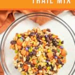 A clear bowl filled with a colorful Halloween trail mix, featuring candies, nuts, and dried fruits. An orange banner at the top reads "Easy Recipe Halloween Trail Mix.