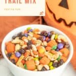 A white bowl filled with Halloween-themed trail mix, featuring candy corn, colored candies, nuts, and raisins. Text reads "Easy Halloween Trail Mix - Festive Fun" with a carved pumpkin in the background.