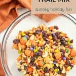 A glass bowl filled with halloween trail mix—nuts, candy corn, raisins, and festive candies—sits on a table adorned with autumn decorations.
