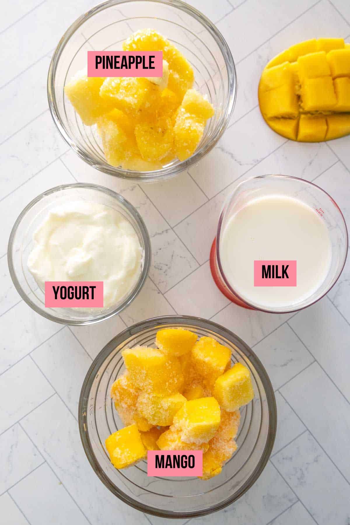 Overhead view of bowls containing labelled ingredients: pineapple, mango, yogurt, and milk, placed on a white tiled surface.