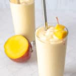 Two glasses of mango smoothie garnished with mango pieces. A stainless steel straw in each glass. A fresh mango half is in the background on a white tiled surface.