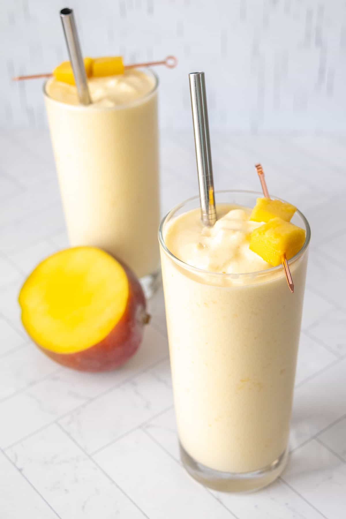 Two glasses of mango smoothie garnished with mango pieces. A stainless steel straw in each glass. A fresh mango half is in the background on a white tiled surface.