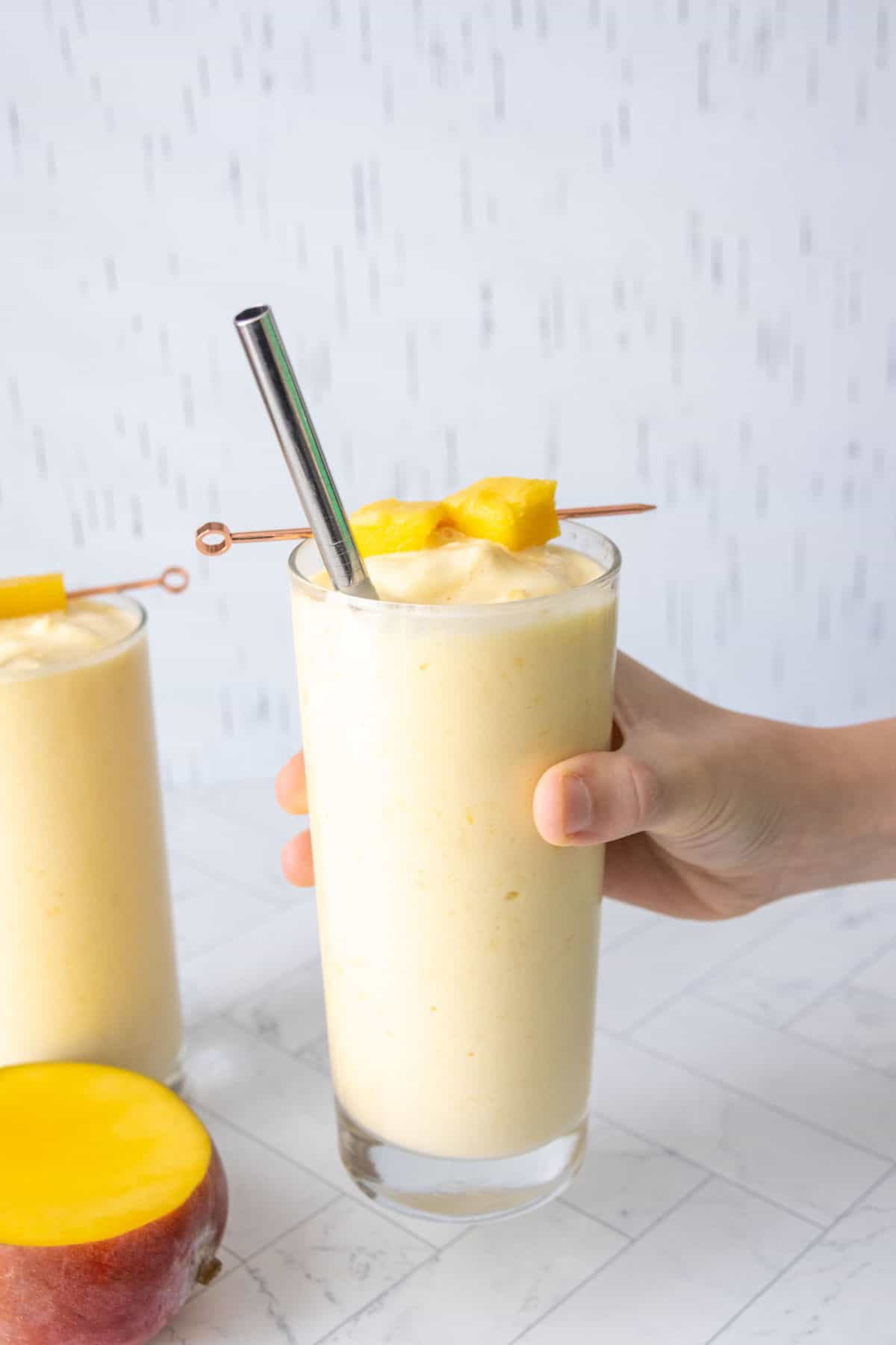 Hand holding a tall glass of yellow mango smoothie with a metal straw and garnished with mango chunks. Another glass of smoothie and a sliced mango are visible in the background.