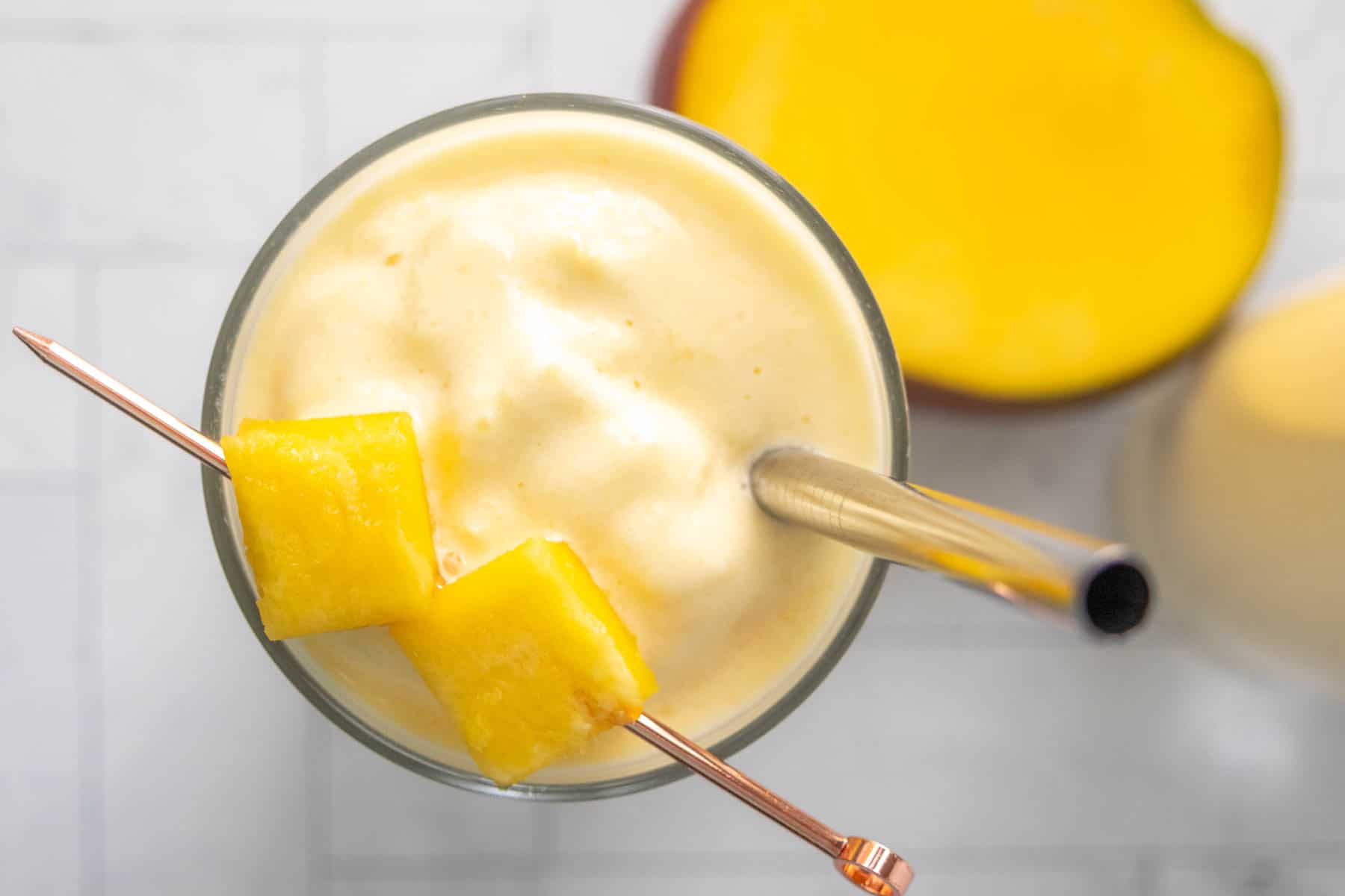 Close-up of a mango smoothie in a glass with a metal straw and two mango chunks on a skewer, placed on a light-colored surface.