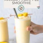 A hand holds a glass of mango pineapple smoothie with a metal straw and fruit garnish; another mango pineapple smoothie and a sliced mango are in the background.
