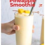 A hand holds a glass of mango pineapple smoothie garnished with pineapple chunks. The text "Mango Pineapple Smoothie" is displayed above the glass. The website "happysnackcidents.com" is noted below, ensuring you don't miss out on this tropical delight.