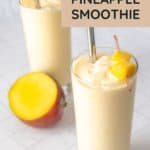 Two glasses of mango pineapple smoothie with metal straws, garnished with fresh mango chunks; a half mango rests beside the vibrant smoothies.