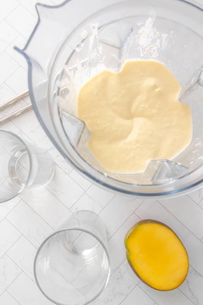 A blender jar with a creamy yellow smoothie mixture inside is on a white tiled surface. Beside it are two empty glasses and a halved mango.