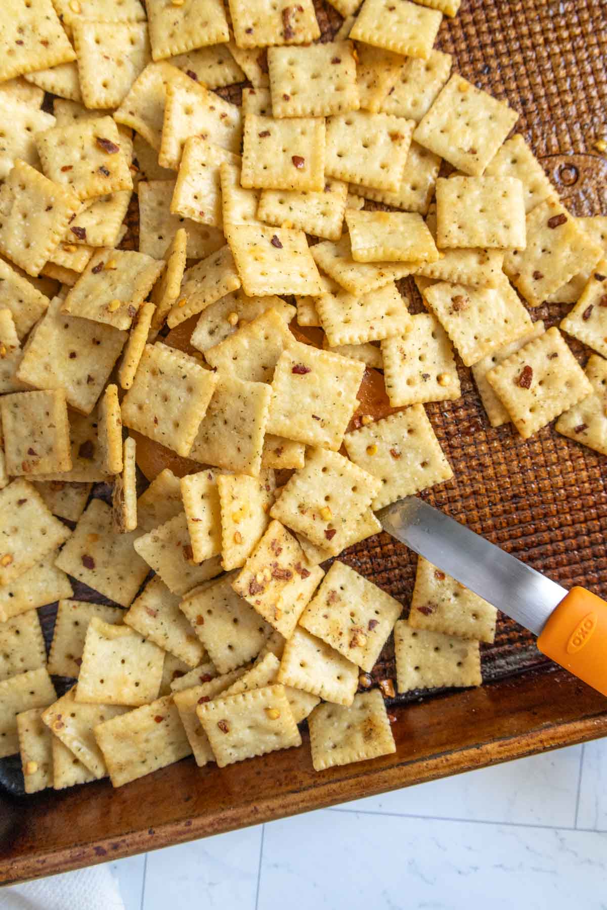 A baking sheet filled with square crackers seasoned with visible spices. A paring knife with an orange handle rests on the edge of the sheet.