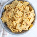 A bowl filled with seasoned square crackers, placed on a light-colored surface with a cloth nearby.