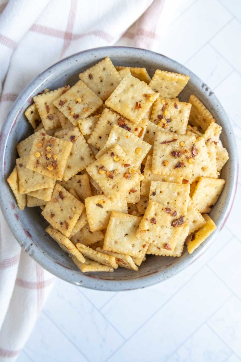 A bowl filled with seasoned square crackers, placed on a light-colored surface with a cloth nearby.