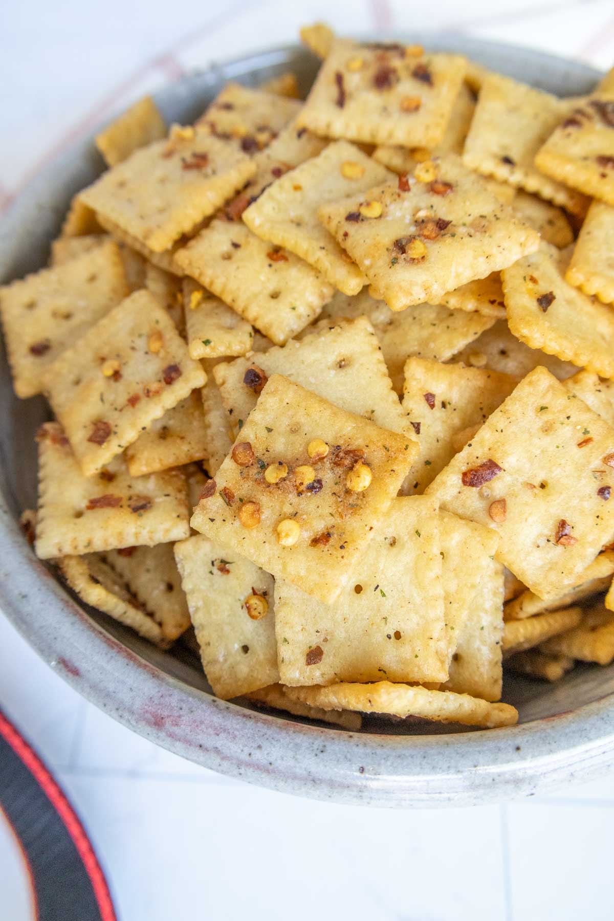 A bowl filled with seasoned square crackers, topped with visible spices and herbs.