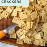 A baking tray filled with square, seasoned firecracker crackers labeled "Spicy & Tangy Firecracker Crackers," with a spatula partially visible in the bottom left corner.