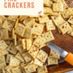 A baking sheet filled with seasoned square crackers. A small knife is resting among the crackers. Text overlay reads "Easy Recipe Alabama Fire Crackers.