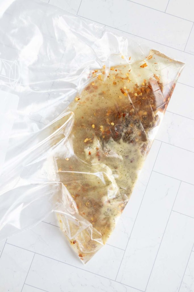 A clear plastic zip-lock bag containing marinated meat and visible spices, placed on a tiled surface.