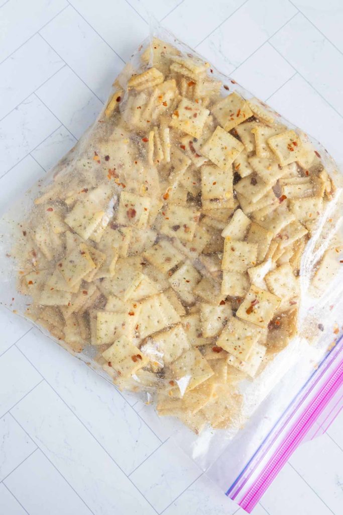 A plastic bag filled with seasoned crackers, featuring specks of red pepper flakes, placed on a tiled surface.