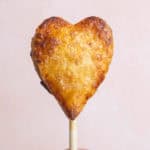 A heart-shaped pastry on a stick held by a hand against a plain background.