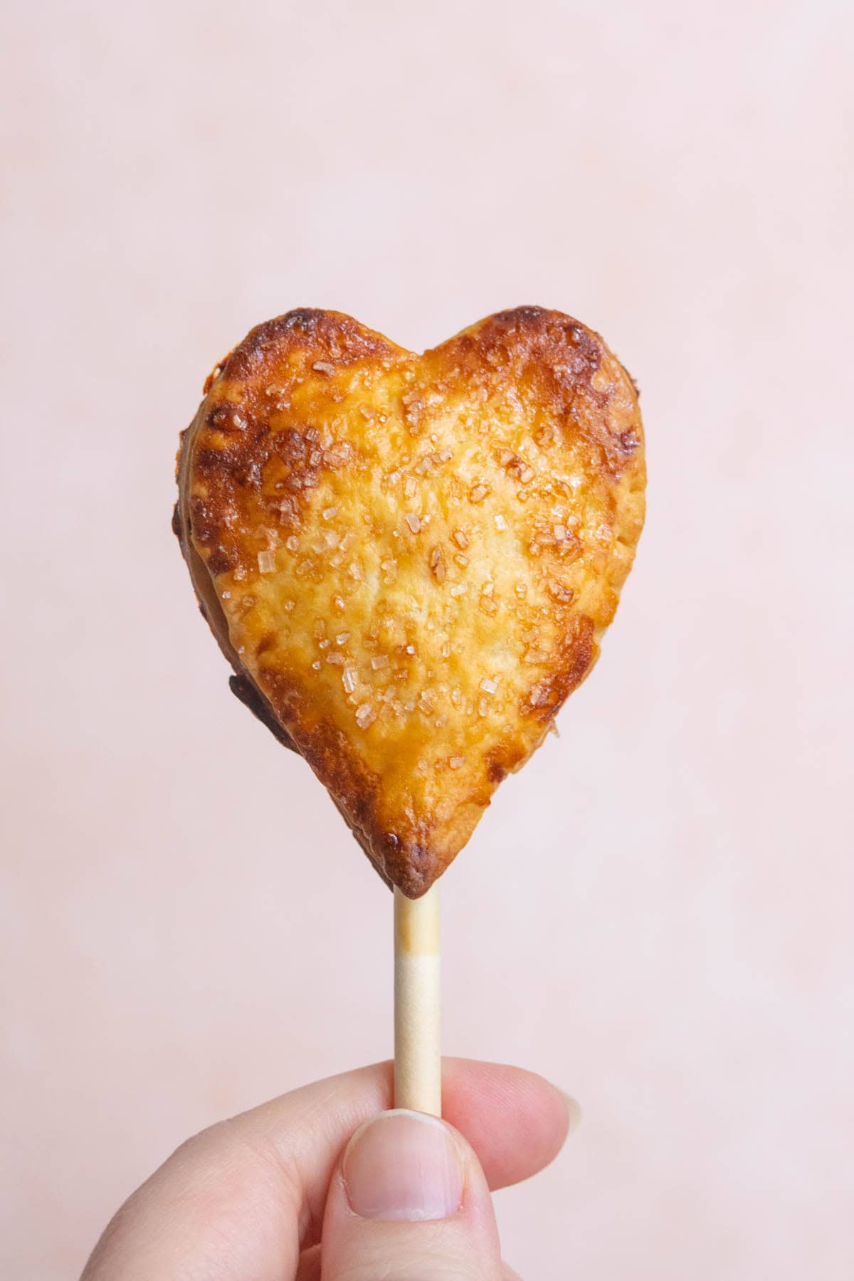 A heart-shaped pastry on a stick held by a hand against a plain background.