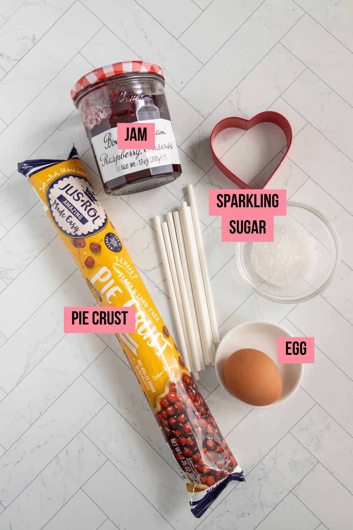 Ingredients for a pie on a white surface: raspberry jam, sparkling sugar, pie crust, one egg, cake pop sticks, and a heart-shaped cookie cutter.