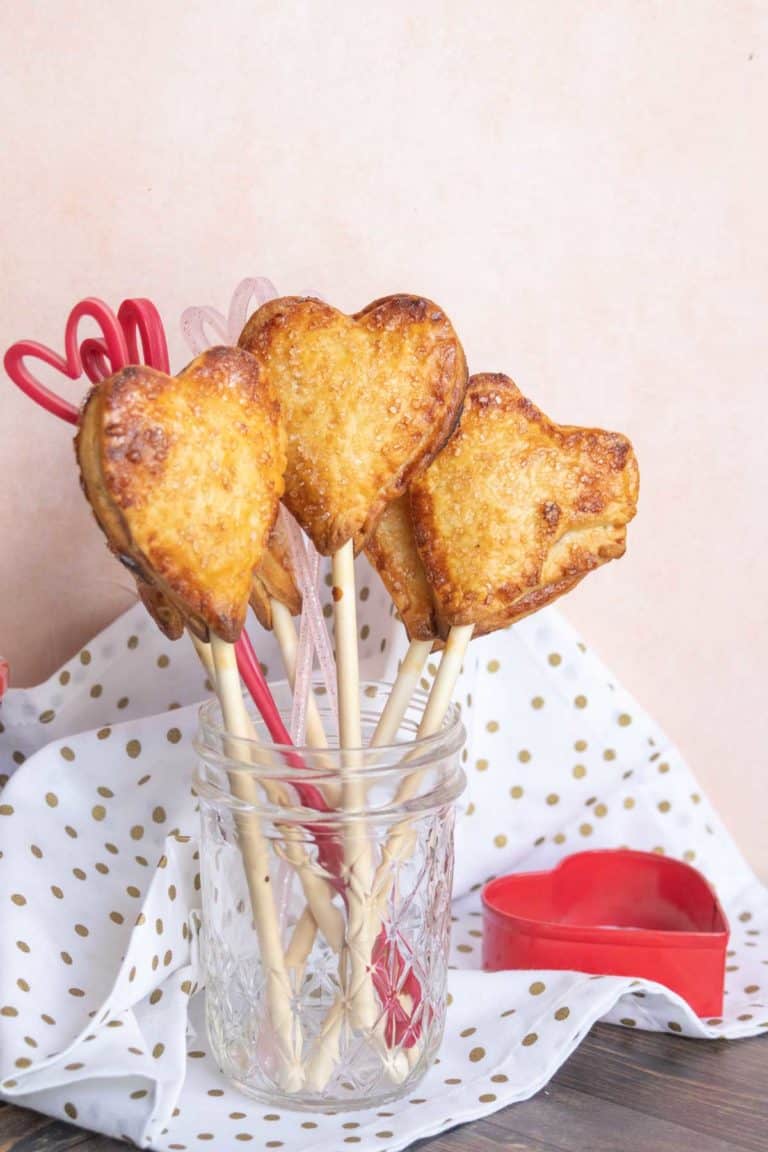 Heart-shaped baked treats on sticks in a glass jar, set on a polka dot cloth. Scissors and a heart-shaped cutter are beside the jar.