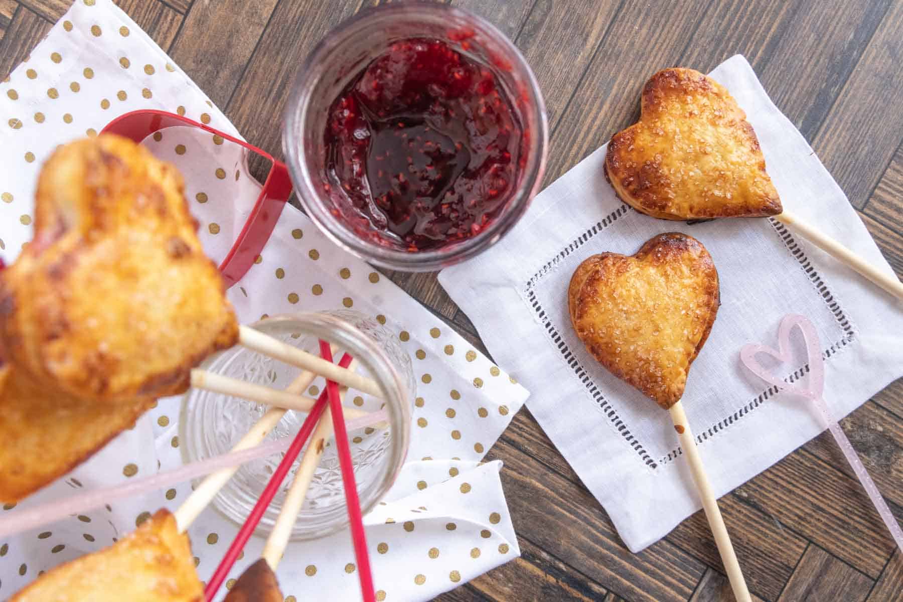 Heart-shaped pastries on sticks are placed on a napkin with a small heart design, next to an open jar of red jam, on a wooden surface.