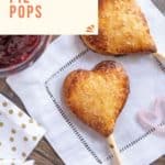 Two heart-shaped jam pie pops on sticks, placed on a wooden surface and white napkin, with a bowl of jam partially visible.