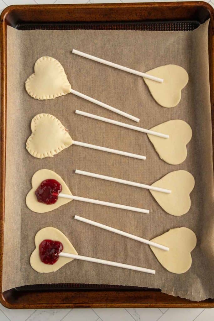 Heart-shaped pastries on sticks, some with red filling, arranged on a baking sheet lined with parchment paper.