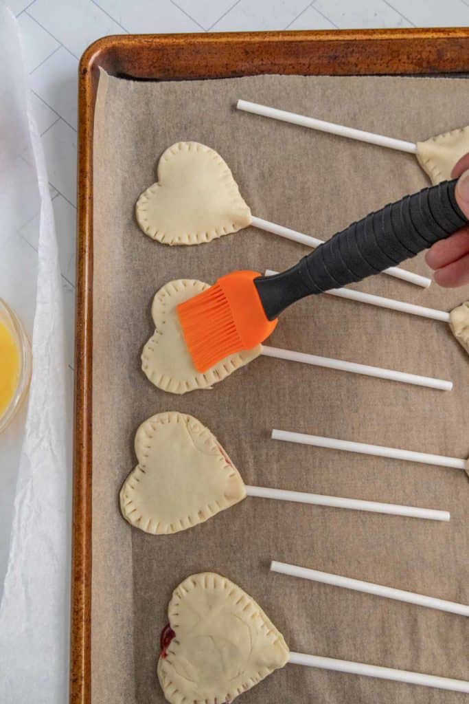 A hand uses an orange brush to apply egg wash to heart-shaped pastry lollipops on a baking sheet lined with parchment paper.