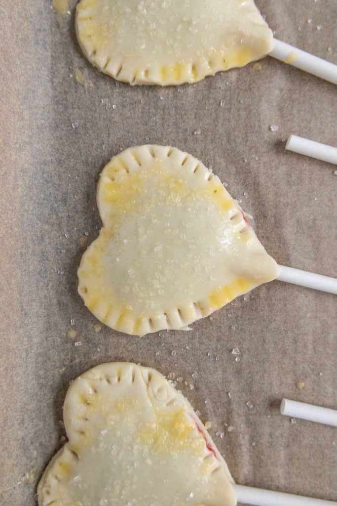 Heart-shaped pie pops on sticks, with crimped edges, sprinkled with sugar, placed on parchment paper.