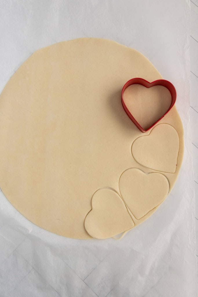 Round sheet of dough with three heart-shaped pieces cut out using a red heart-shaped cookie cutter.