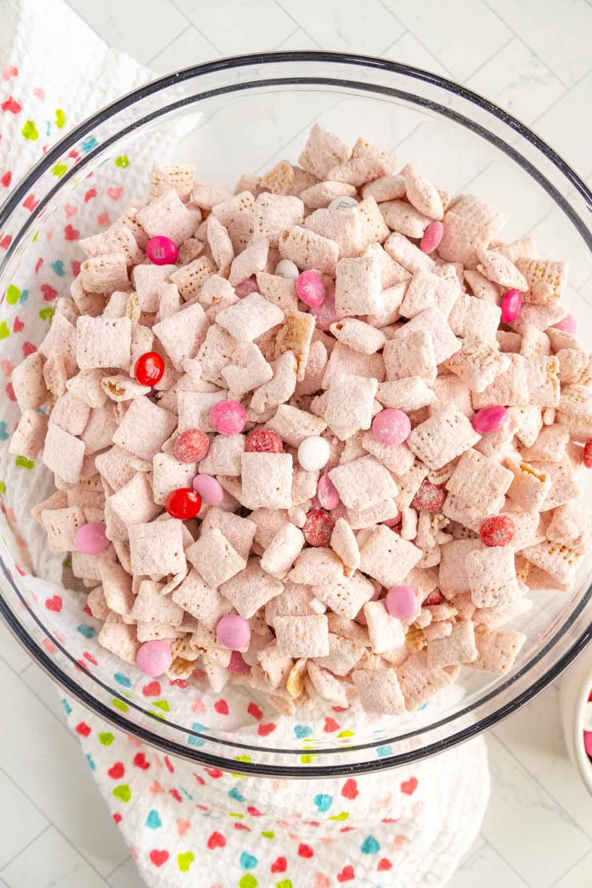 A glass bowl filled with Chex cereal coated in powdered sugar, mixed with pink and red candy-coated chocolates. A printed fabric is partially visible underneath.