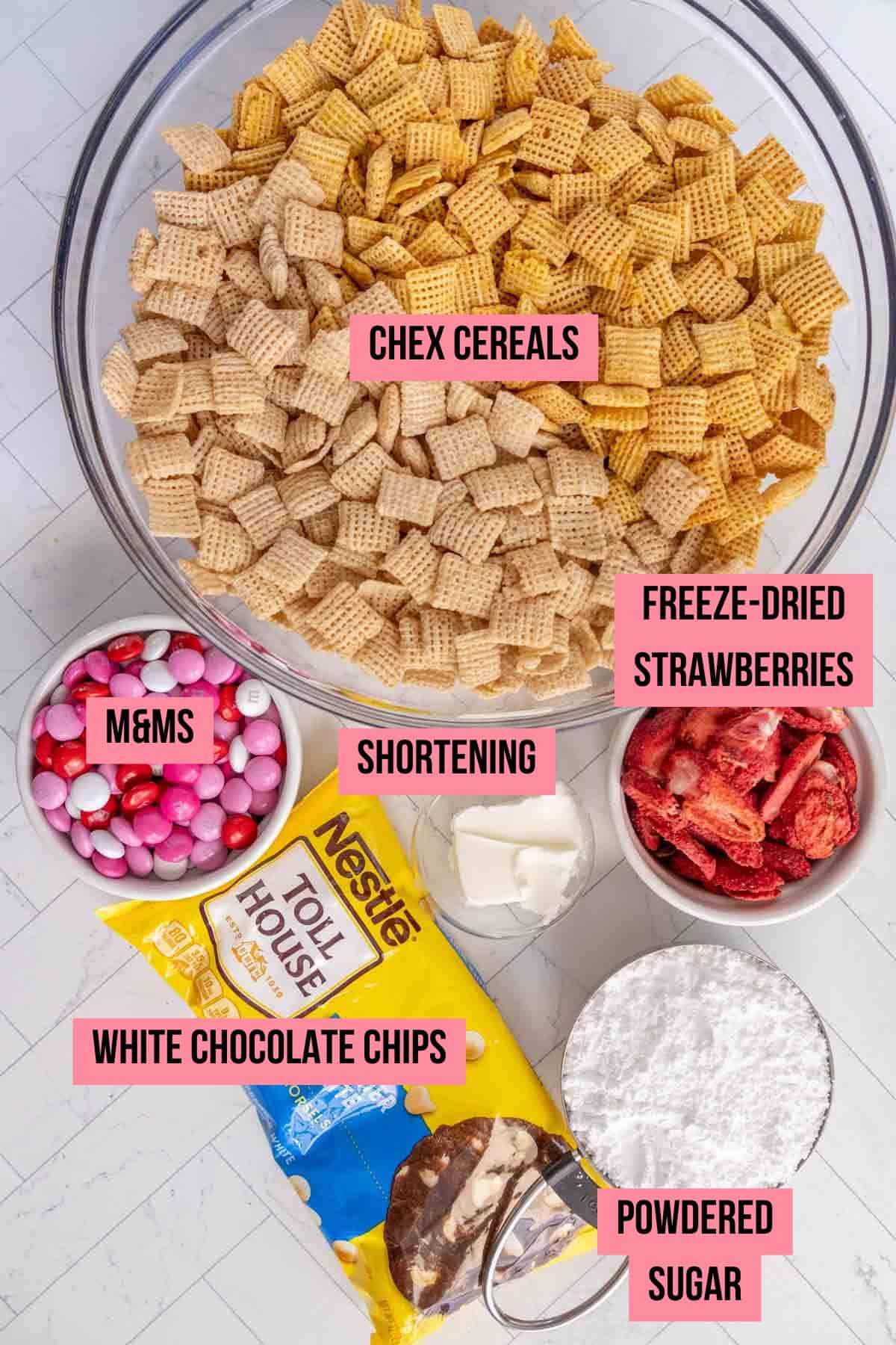 Ingredients for a snack mix: Chex cereals in a large bowl, freeze-dried strawberries, M&Ms, shortening, white chocolate chips, and powdered sugar, all arranged on a countertop.