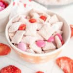 A bowl of powdered cereal mix with pink and red M&Ms, surrounded by dried strawberry slices on a white surface.