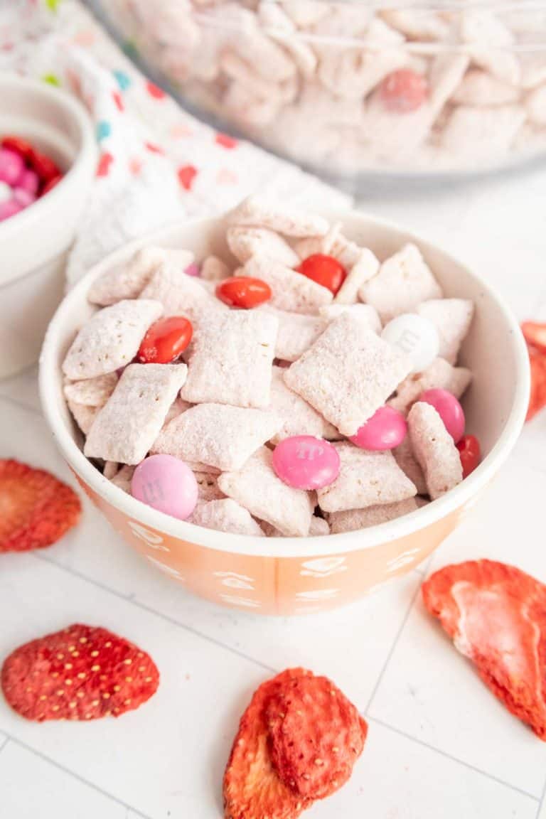 A bowl of powdered cereal mix with pink and red M&Ms, surrounded by dried strawberry slices on a white surface.