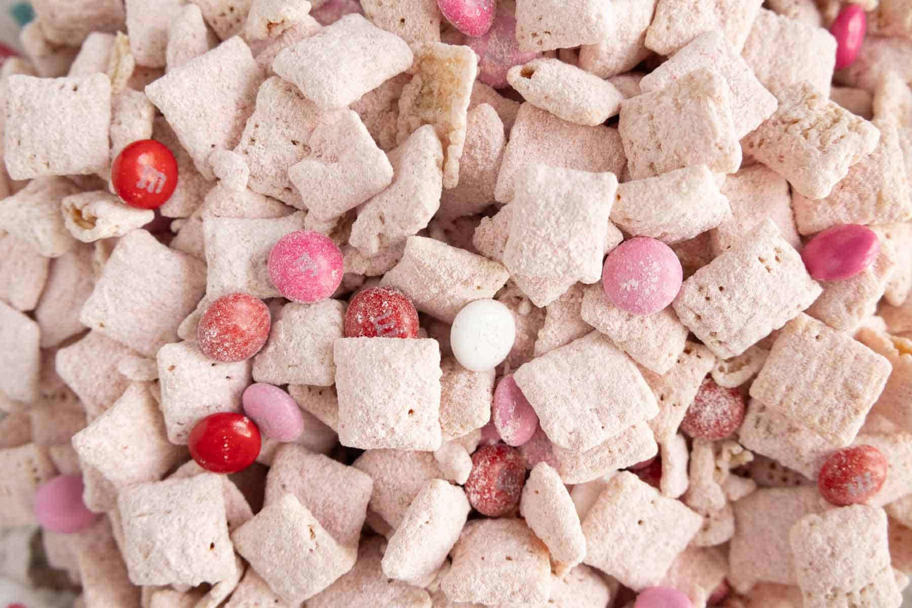 Close-up of a snack mix with cereal squares covered in powdered sugar, interspersed with pink, red, and white candy pieces.